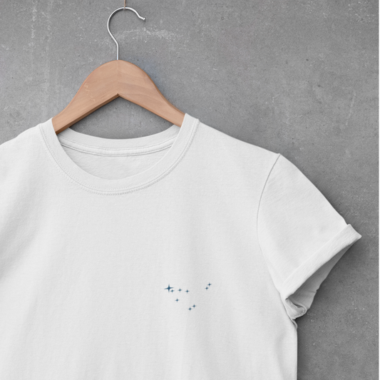 White Capricorn constellation t-shirt on a wooden hanger against a gray concrete wall.